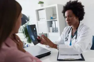 A woman wearing a white lab coat is holding a scan and showing it to a woman wearing a pink shirt. They are both sat down in a room with a clipboard of papers in between them.