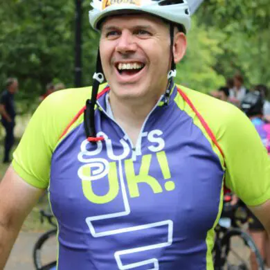 Stuart is smiling widely at the camera. He is wearing the Guts UK technical t-shirt and a white helmet. He is outdoors with green trees and bikes behind him.