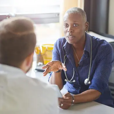 A female doctor sits at her desk and chats to a male patient while looking at his test results on her digital tablet . She is a blue shirt with the sleeves rolled up and a stethoscope around her neck.