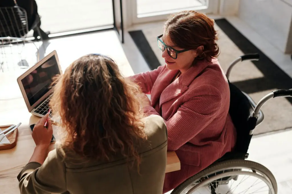 A cropped photo of two women sat chatting in front of a laptop. One woman is in a wheelchair and wears a red jacket and the other wears a khaki blouse.