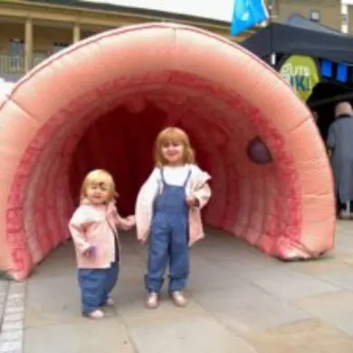 Two young girls are standing outdoors in front of Colin, the inflatable Colin. They are smiling at the camera and holding hands. They are wearing the same outfit: a blue denim dungaree with a white T-shirt underneath and a pink jacket.
