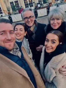 Geoff is stood in the centre on the street in front of a restaurant. He is smiling wearing his black trench coat and coloured pattern scarf. He is stood beside 4 of his family members all smiling.