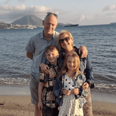 Sarah is standing on the beach with the sea waves behind her. She is standing next to her husband with his hand on her shoulder. She has her hands wrapped around her two children who are standing in front of her. All four of them are smiling at the camera.