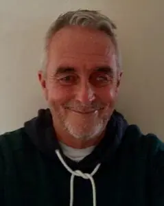 Geoff is taking a selfie and smiling at the camera at home. He is wearing a black hoodie. 