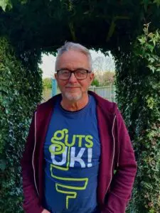 Geoff is wearing the Guts UK blue t-shirt with a burgundy jacket on top. He is stood smiling at the camera stood under a bushy garden hedge.