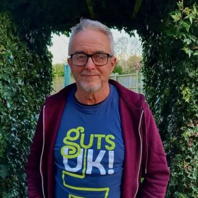 Geoff is wearing the Guts UK blue t-shirt with a burgundy jacket on top. He is stood smiling at the camera stood under a bushy garden hedge.