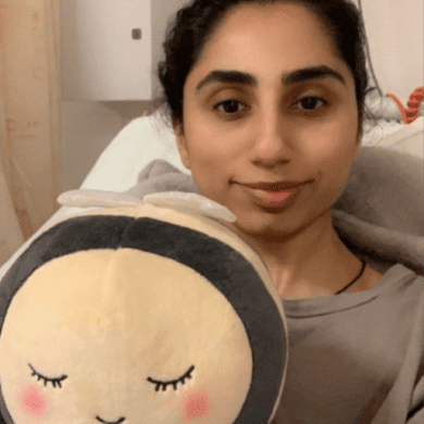 Mia is taking a selfie, sitting in a hospital bed. The selfie is cropped to show from the top of her head down to just below her chest. She is smiling slightly at the camera and holds a cuddly bee towards the camera.