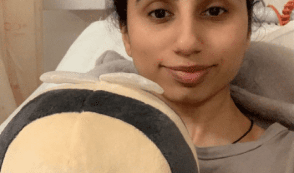 Mia is taking a selfie, sitting in a hospital bed. The selfie is cropped to show from the top of her head down to just below her chest. She is smiling slightly at the camera and holds a cuddly bee towards the camera.