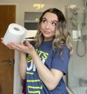 Abbie is standing in a white tiled bathroom. She is smiling facing the camera with her eyes staring to her right. She is wearing the navy Guts UK T-shirt and holding a toilet roll with both her hands.