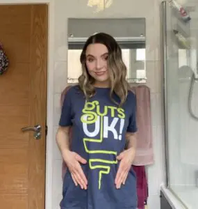 Abbie is standing in a bathroom with a mirror and towels hanging behind her. She is smiling at the camera and wearing the navy Guts UK T-shirt with her hands placed on her stomach.