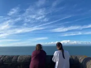 A mother and her adult daughter stand together with their backs to the camera. They are looking out onto the sea and you can see the blue sky above.