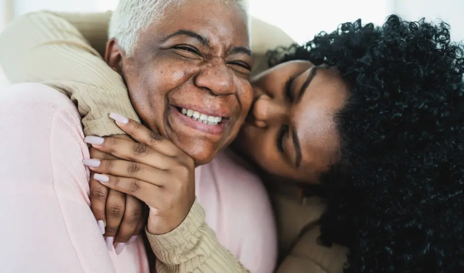 A mother is smiling as her adult daugher hugs her and gives her a kiss on the cheek.