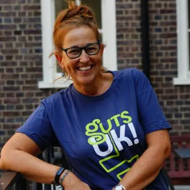 Suzanne is outside, she is standing up with her arm leaning on a railing. She is wearing a Guts UK branded t-shirt and glasses. Her hair is tied up. She is smiling at the camera.