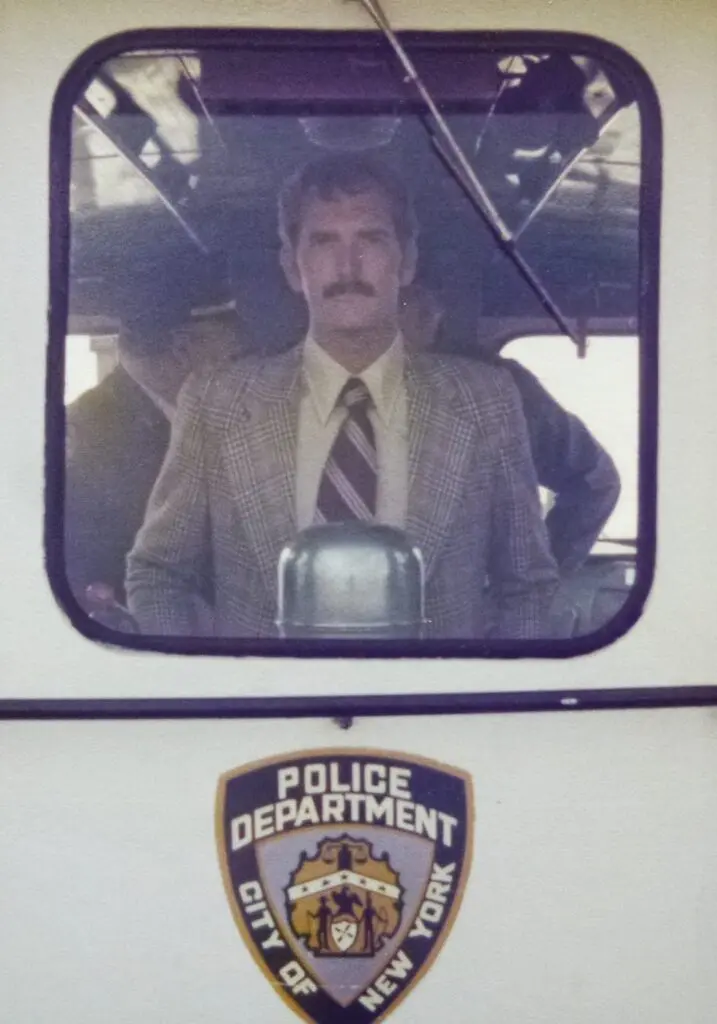 Tony is on a police boat during his career in the police force in Bermuda. The photo is taken through the window and he is looking at the camera with a neutral expression.