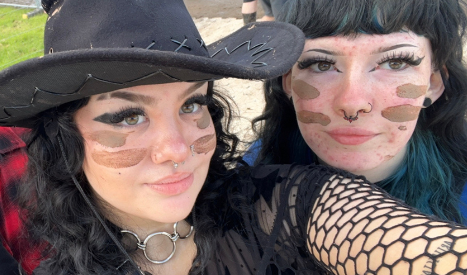 Rachel (left) wears a black cowboy hat, with fishnet style sleeves and a top and has black, long, curly hair. She is smiling at the camera and has two stripes of brown face paint on each of her cheeks. She is with her friend at a music festival.