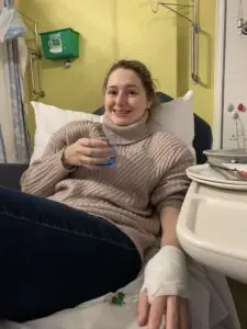 Sian is wearing a hazelnut jumper with black leggings and smiling at the camera. She is sat on a hospital bed with her left arm bandaged up and holding a glass of water on her right hand.