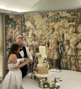 Sian is wearing a white wedding dress and Matt in a tuxedo during their wedding. They are holding a cake knife together, smiling while cutting their wedding cake. Renaissance art is seen on the walls behind them.