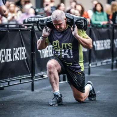 Cornelius has weights on his back and lunged forward. He is wearing a Guts UK t-shirt with black shorts and trainers while completing a race.