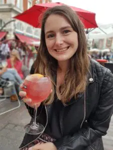 Sian is smiling at the camera in her black jacket. She is sat down at and holding a pink cocktail.