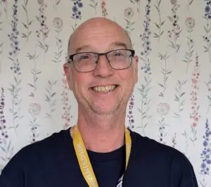 Stephen stands in front of a white wallpaper background, with delicate, floral detailing on it in different colours. It is a headshot of Stephen, and he wears black, rectangular glasses with curved edges, a black t-shirt and a yellow lanyard. He is smiling at the camera.