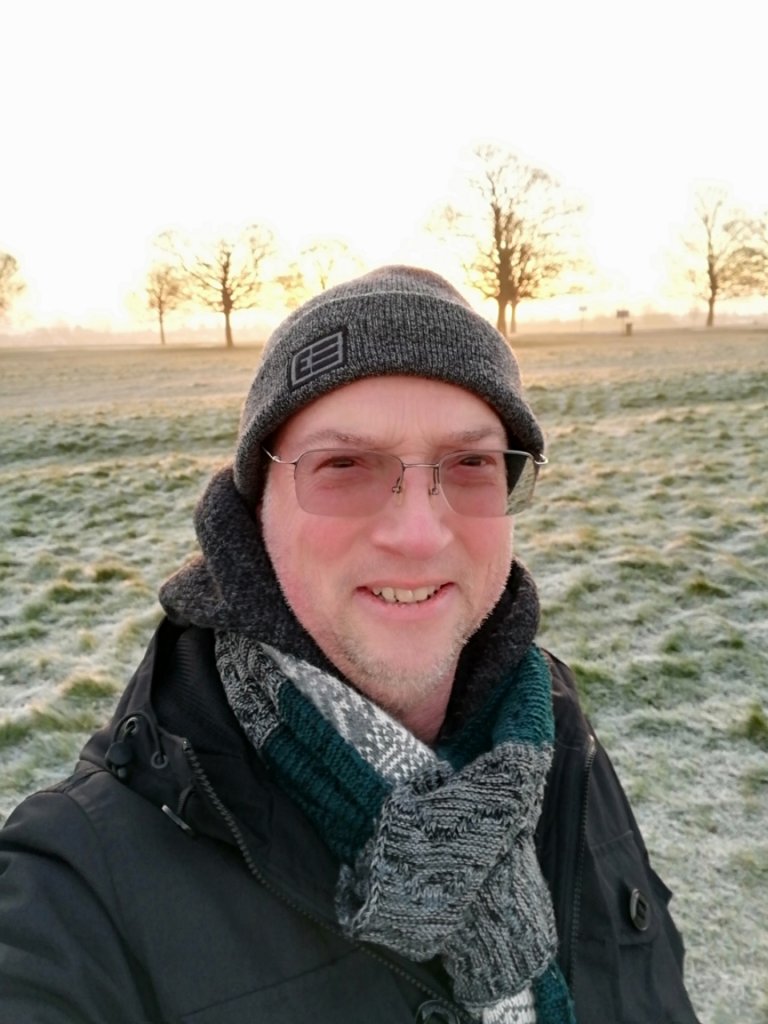 Stephen is taking a selfie outdoors, with a frosty, grassy background and the outlines of trees in the background in the sun. He is wearing a grey beanie hat, a black coat and has a green, grey and white knitted scarf on. He is smiling and wears black framed, rectangular glasses, with curved edges.