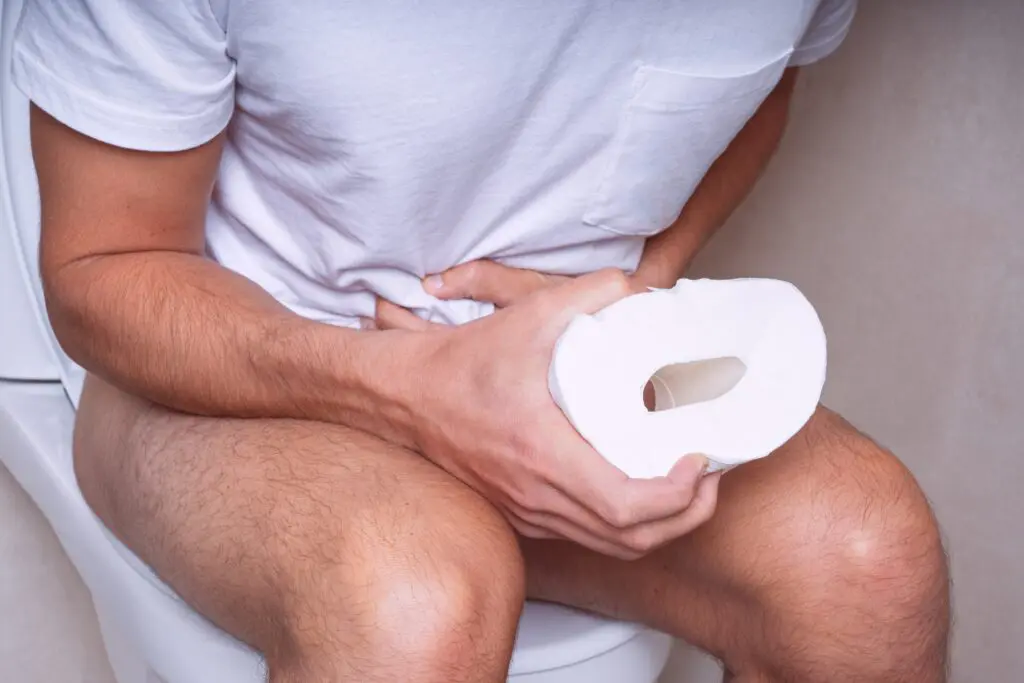 Man sitting on the toilet, clutching his abdomen and squeezing a toilet roll between his other hand.