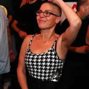 Cheryle is dancing with one arm above her head and is smiling. She wears black rimmed glasses and a black and white dogtooth vest top. She has a bottle of water tucked in to the top of her black trousers, which you can see the waist of.