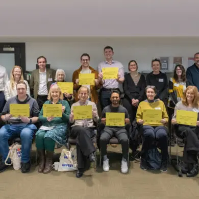 The photograph shows a group of 20 people standing and sitting together. They are smiling at the camera. Some of them are holding up pieces of paper with the top ten research priorities for diverticular disease on them.