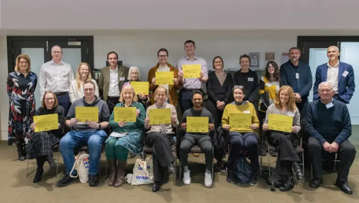 The photograph shows a group of 20 people standing and sitting together. They are smiling at the camera. Some of them are holding up pieces of paper with the top ten research priorities for diverticular disease on them.