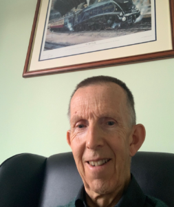 Dave is sat on a black armchair. He is smiling at the camera. He has short dark hair and is wearing a shirt with a collar. The wall behind him is light green and there's a framed picture of a train on the wall behind him.
