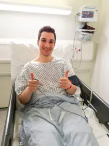 Ziyad is sitting up in a hospital bed, wearing a hospital gown. A medical monitor is beside him. He’s smiling at the camera with both thumbs up.