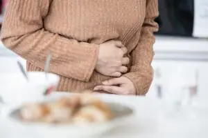 A person's torso is seen, covered by a light brown jumper. They arms are covering their stomach, with their hands gripping their upper abdomen, indicating pain or discomfort.