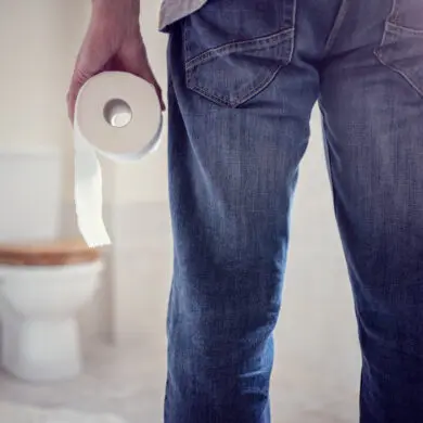 The image shows a person's legs, standing in front of a toilet. They are wearing blue jeans. They are holding a role of toilet paper in their hands.