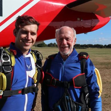 William on the right in his blue sky diving suit and gear standing next to his instructor both smiling at the camera. They are stood under a plane's wing in a plain field.
