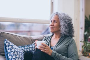 A woman with curly grey hair is sat on a light coloured sofa with blue patterned cushions. She is holding a white mug as she looks out of the window, smiling. The view fro the window is blurred.