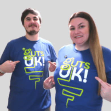 Wayne is standing on the left and Jessica is standing on the right against a light grey background, wearing Guts UK t-shirts. They are facing the camera, smiling and pointing to the Guts UK logo on their shirts.