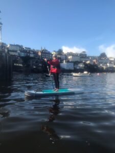 Sarah is wearing a wetsuit and a red life jacket on a still lake standing on a paddleboard, looking towards the camera