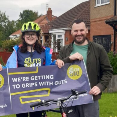 A woman and man is stood in front of a house and they are holding the Guts UK banner together. The woman is wearing a green helmet and both are smiling at the camera.