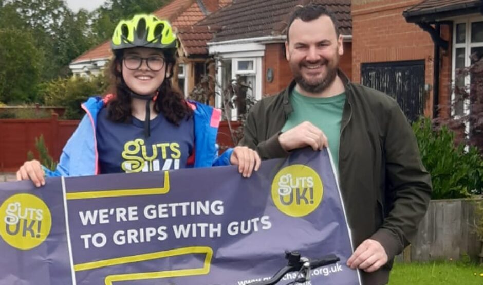 A woman and man is stood in front of a house and they are holding the Guts UK banner together. The woman is wearing a green helmet and both are smiling at the camera.