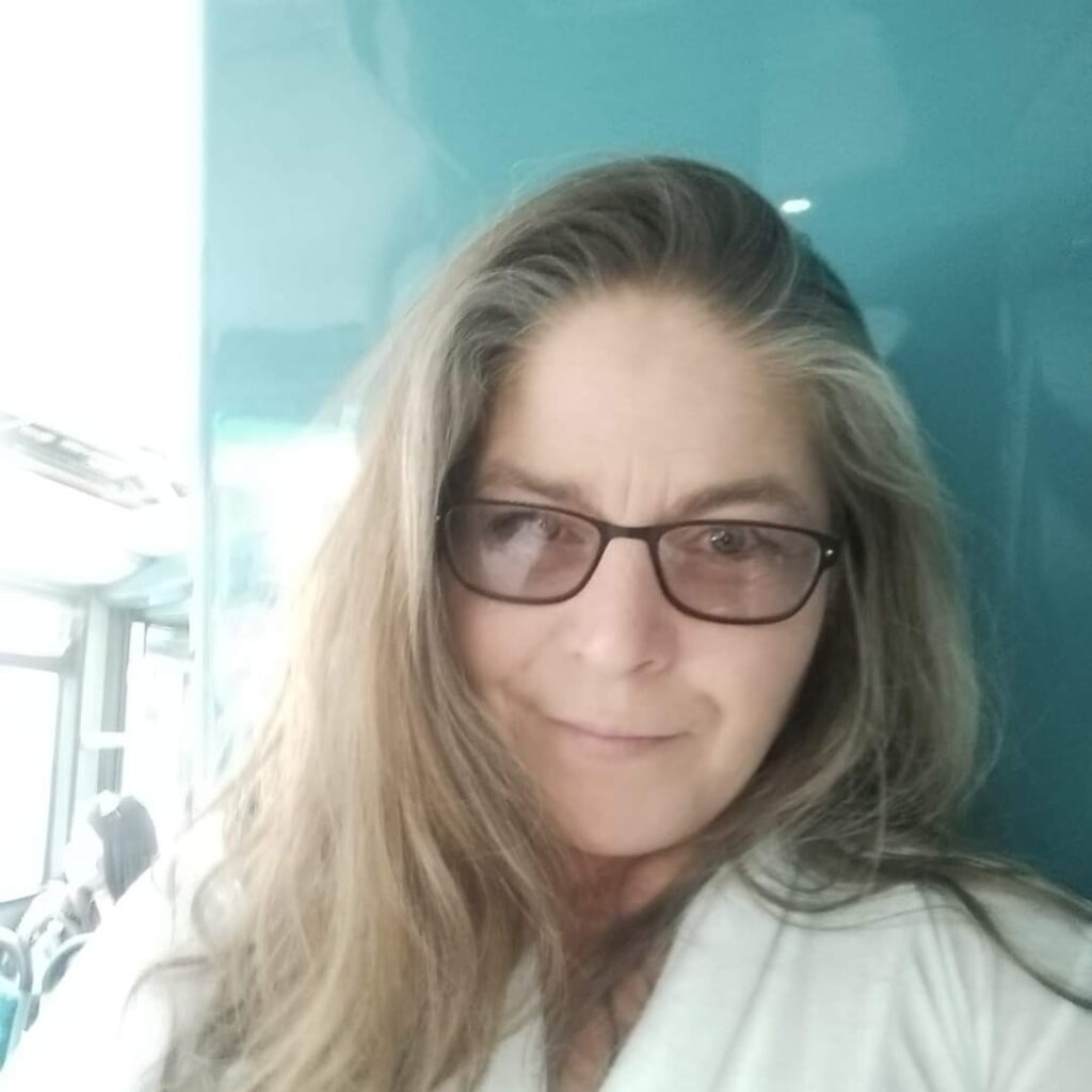 Cheryle is smiling at the camera and is wearing black-framed glasses. She is taking a selfie and the photo is cropped to show from just below her shoulders, upwards.