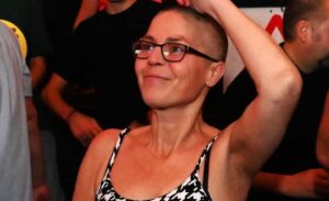 Cheryle is at a rave, dancing, with one arm up in the air. She is smiling and wears a dogtooth print vest top and black framed glasses. The photo is cropped from her waist upwards.