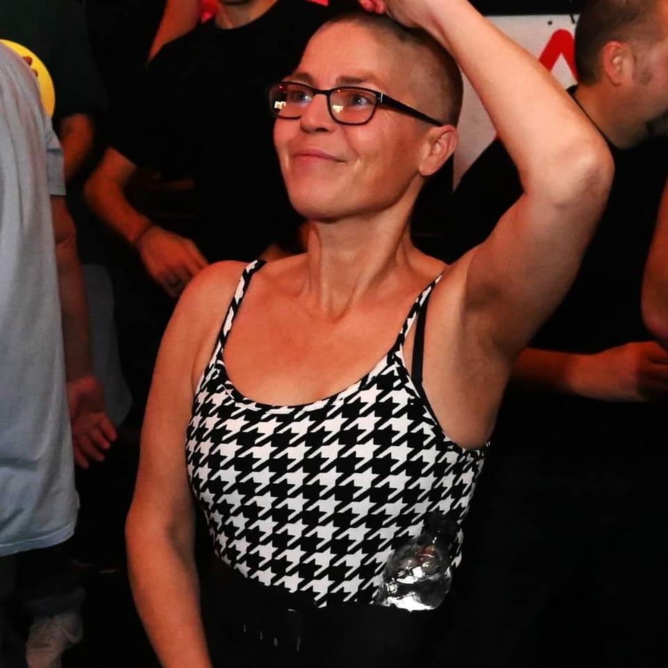 Cheryle is at a rave, dancing, with one arm up in the air. She is smiling and wears a dogtooth print vest top and black framed glasses. The photo is cropped from her waist upwards.