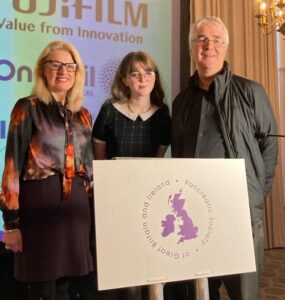 Lola stands at the speech podium, with her dad John on her right and Guts UK's Patient and Public Involvement and Engagement Officer, Helen to her left. They are all looking at the camera and smiling.