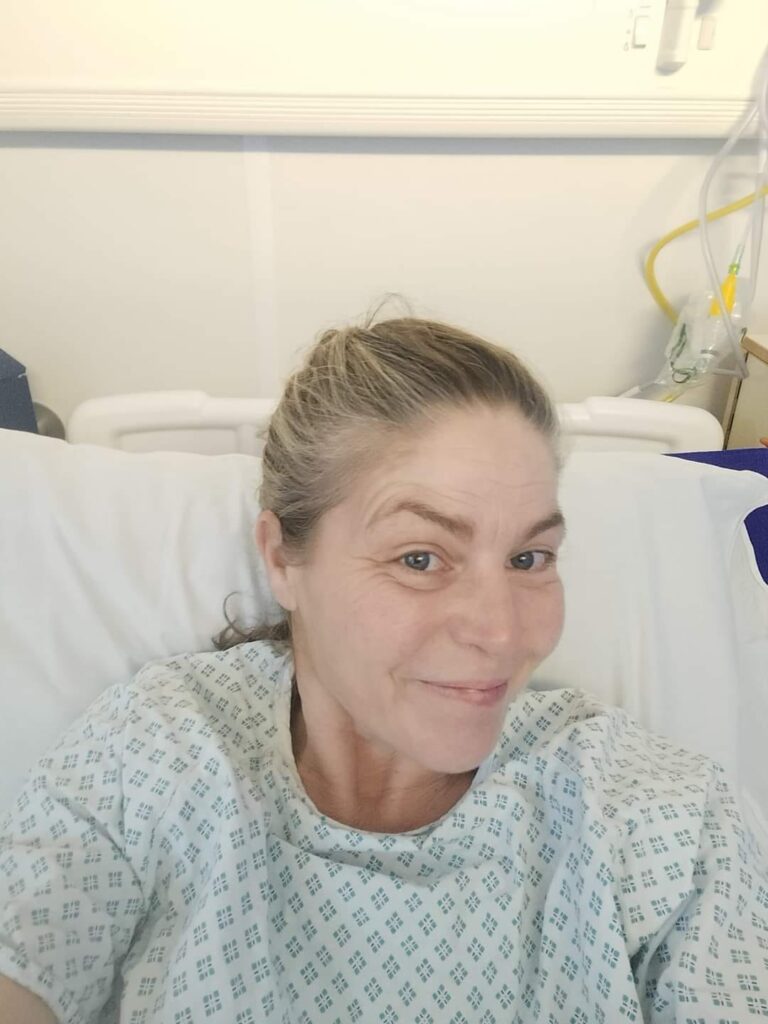 Cheryle is taking a selfie. She is smiling and is sat in a hospital bed, wearing a white hospital gown with small green diamonds printed on it. The photo is cropped to show just above her chest, upwards.