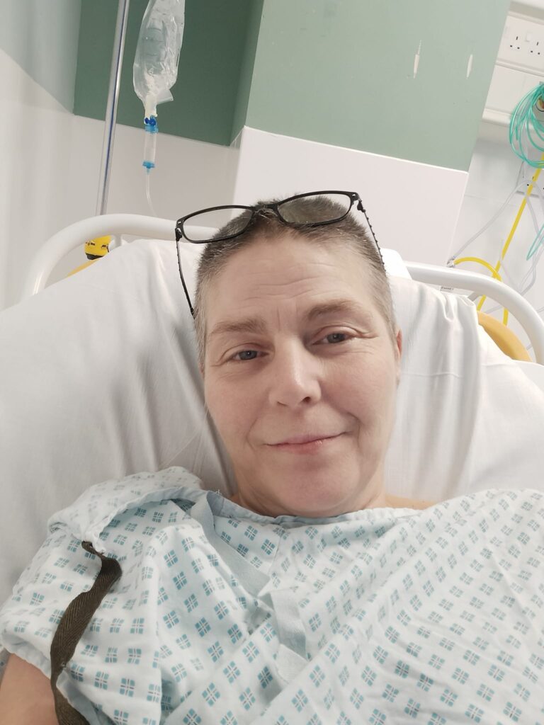 Cheryle is taking a selfie in a hospital bed. She is wearing a white hospital gown with small, green diamonds printed on it. Her glasses are propped up on her head and she is smiling at the camera. The photo is cropped to show above her chest upwards.