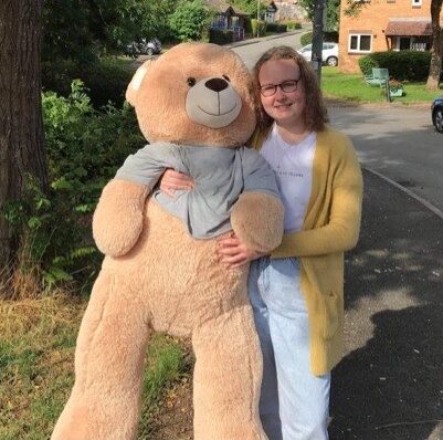 Dominika is wearing a white top and light denim trousers and a yellow cardigan. She is stood outside, holding a pale brown giant teddy bear and she is smiling at the camera. She wears black rimmed glasses.