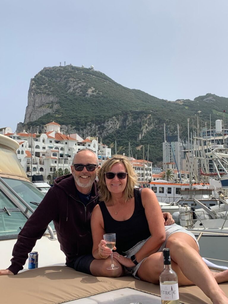 Mark and Jules are sitting together on a boat, smiling at the camera. Jules is holding a glass of white wine and they both are wearing black sunglasses, black tops and shorts.