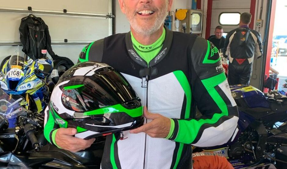 Mark is standing in a garage. He is wearing black, green and white motorbike leathers and is holding a matching helmet. He is smiling at the camera and wears black sunglasses. There is a motorbike behind him.