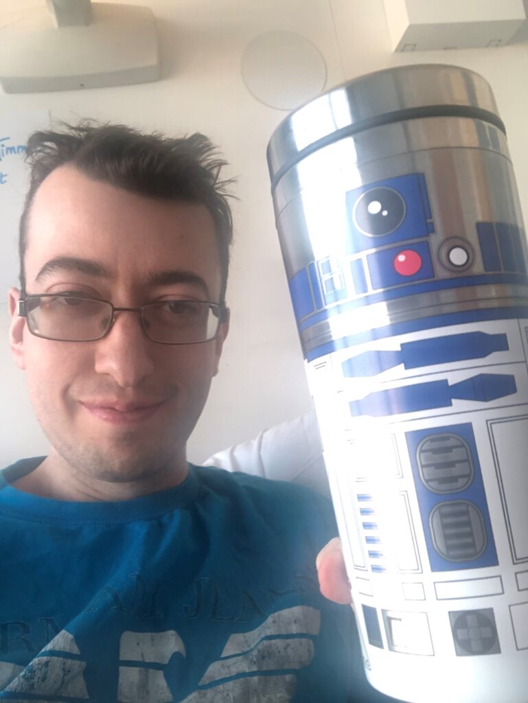 Matthew is taking a selfie, from his chest upwards and is holding a insulated R2D2 Star Wars beaker up to the camera, smiling. He is in a hospital bed and wears black rimmed curved, rectangular glasses, and a blue t-shirt. His hair is short and is gelled up into spikes.
