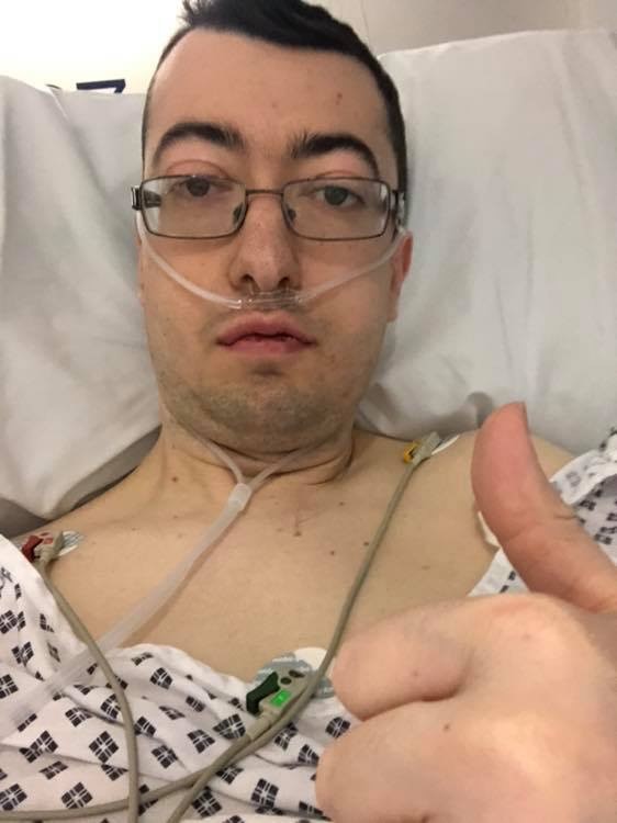 Matthew is taking a selfie, from his chest upwards and is holding his thumb up at the camera. He is in a hospital bed and has a nasal cannula delivering oxygen and is fitted up to various monitors and wires. He wears black rimmed curved, rectangular glasses.